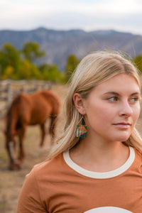 Thumbnail for A blonde woman in a rust-colored t-shirt with a white collar is shown in profile, wearing colorful geometric dangle earrings. The background is a soft-focus outdoor scene with a brown horse grazing in a paddock and mountains in the distance.