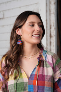 Thumbnail for A woman with long wavy brown hair is shown in a close-up portrait, wearing colorful geometric drop earrings and layered gold necklaces, paired with a multicolor plaid shirt against a white painted brick wall.